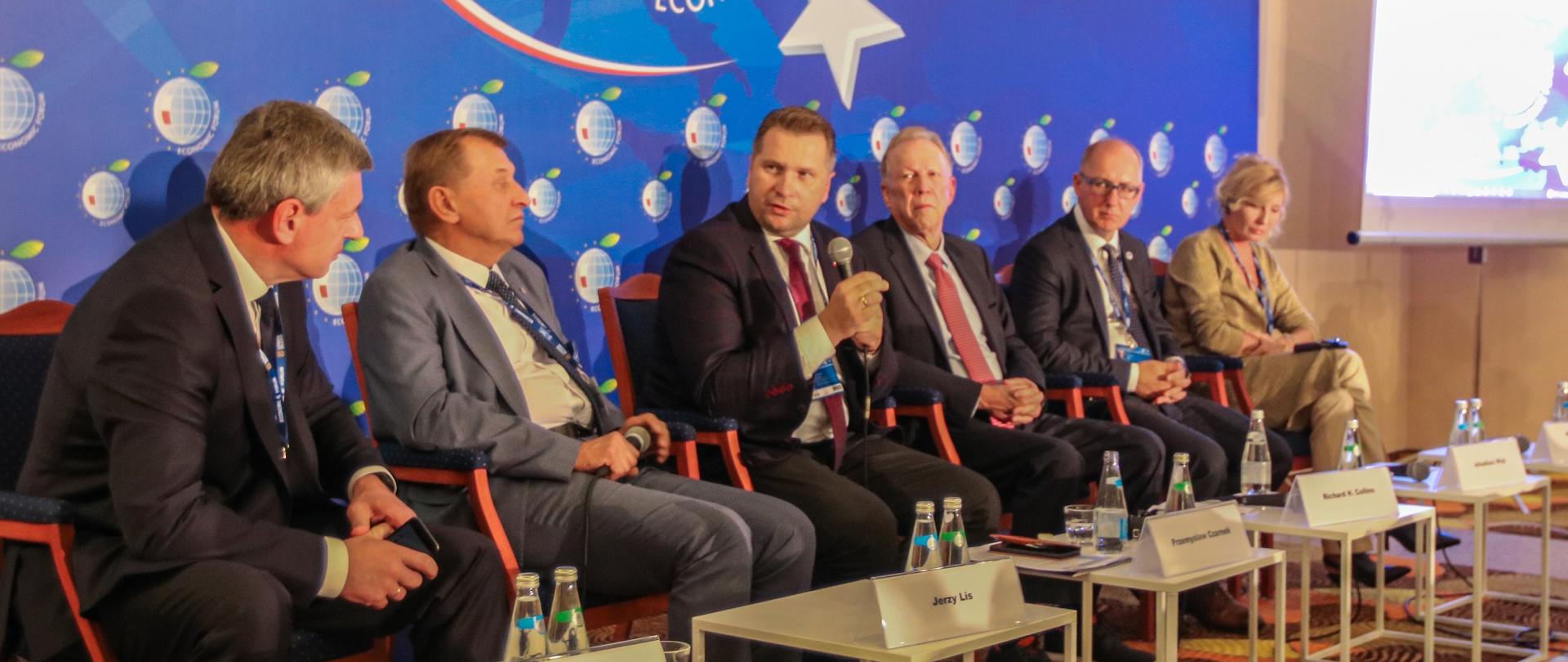 Minister Czarnek i kilku mężczyzn w garniturach siedzą na krzesłach ustawionych w rząd, przed nimi stoliki, za nimi niebieska ściana z napisem Economic Forum.
