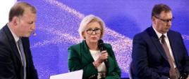 Wiceminister Machałek siedzi na krześle i mówi do trzymanego w reku mikrofonu, po obu jej stronach siedzą mężczyźni w garniturach, jeden trzyma otwartą papierową teczkę. Za nimi fioletowa ściana.