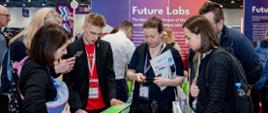 W dużej hali nad stołem na którym leży wiele drobnych elementów nachyla się kilkoro młodych osób, za nimi różowo-fioletowa ścianka z napisem Future Labs.