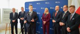 Przed niebieską ścianką stoi grupa mężczyzn w garniturach i jasnowłosa kobieta w ciemnoczerwonym ubraniu, przed nimi minister Czarnek mówi do mikrofonu na stojaku, z tyłu stoi tablica z napisem Branżowe Centra Umiejętności, za nią biała ściana i zasłonięte białymi roletami okno.