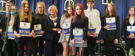 Wiceminister Marzena Machałek z wizytą w Zgorzelcu, wiceminister Marzena Machałek w towarzystwie uczniów Zespołu Szkół Ponadpodstawowych w Zgorzelcu, uczniowie trzymają dokumenty otrzymania stypendium starosty, w tle dekoracja sali.