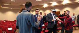 Kilku mężczyzn stoi na środku sali i rozmawia, jeden ma na głowie okulary VR. Za nimi stoją ustawione w rzędy czerwone krzesła.