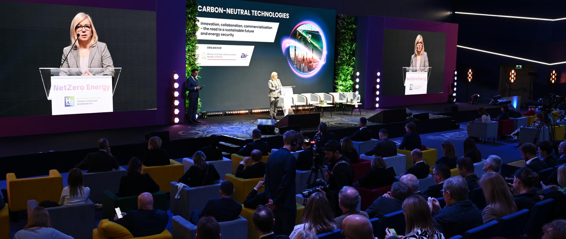 Ministra Klimatu i środowiska Paulina Hennig-Kloska na inauguracji konferencji Ministerstwa Klimatu i Środowiska na terenie Międzynarodowych Targów Poznańskich. Ministra stoi za mównicą, za nią w tle telebim z tytułem konferencji i jej twarzą. 
