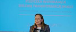 Minister funduszy i polityki regionalnej Katarzyna Pełczyńska-Nałęcz stoi przy pulpicie z mikrofonem i przemawia, za jej plecami grafika informująca o pożyczkach wspierających zieloną transformację miast