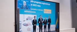 Ekran telewizora na którym widać uczestników konferencji prasowej w Katowicach - w środku przy mikrofonie stoi marszałek województwa śląskiego Jakub Chełstowski