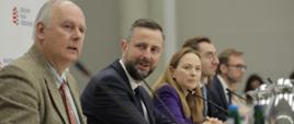 Wiceminister Paweł Zalewski siedzi z lewej strony przy stole i mówi do osób zgromadzonych na sali. 