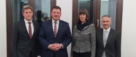 Wiceminister Rzymkowski stoi w towarzystwie dwóch mężczyzn i kobiety za nimi dwa wysokie okna