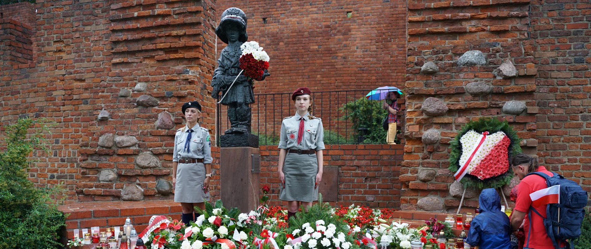 Na zdjęciu widoczny jest wieniec złożony pod Pomnikiem Małego Powstańca. Obok pomnika widać stojące harcerki.