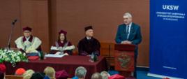 Na sali za mównica stoi wiceminister Murdzek i mówi do mikrofonu, obok niego stół na którym siedzi kilka osób w czerwonych ceremonialnych strojach z gronostajowymi kołnierzami, z boku niebieska ścianka z napisem UKSW.