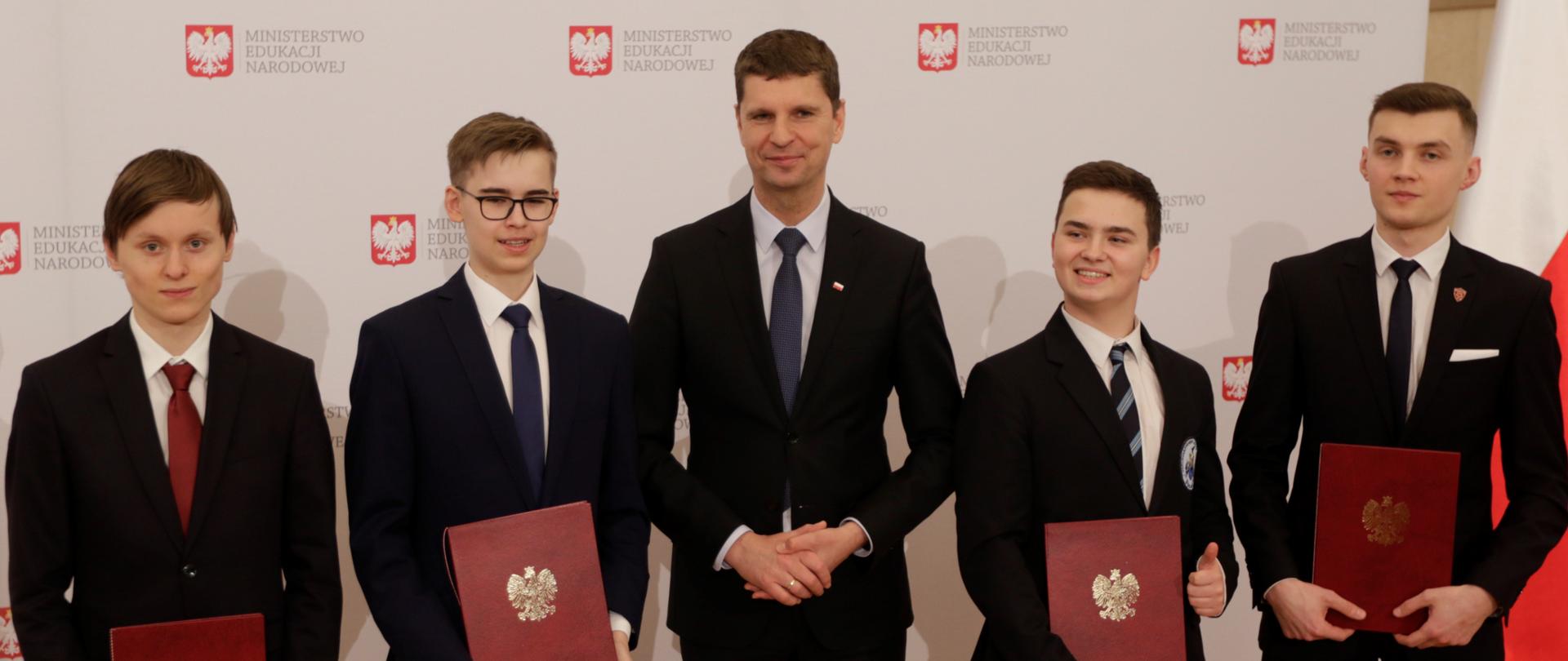 W środku zdjęcia stoi minister edukacji, obok niego uczniowie z otrzymanymi stypendiami. W tyle ścianka z logo Ministerstwa Edukacji Narodowej 