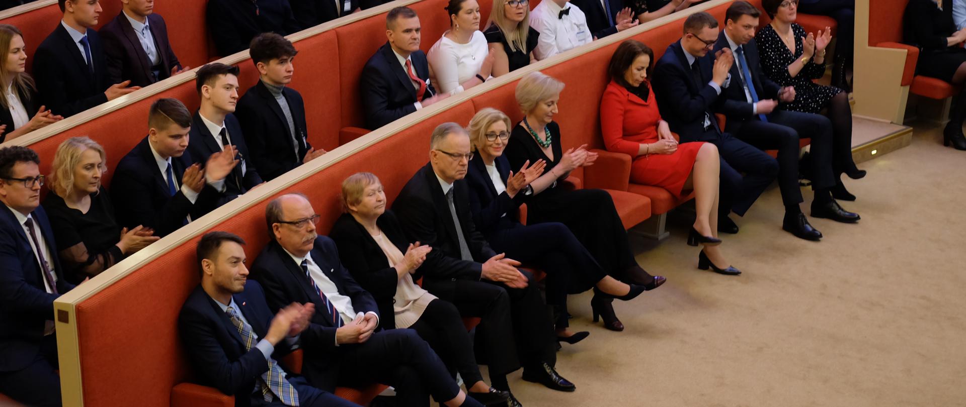 Elegancko ubrani ludzie siedzą w ławach obitych pomarańczowym materiałem i biją brawa. Na środku siedzi wiceminister edukacji narodowej – Marzena Machałek.