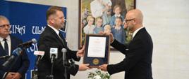 Minister Czarnek stoi pod ścianą na której wisi rodzinny portret, podaje stojącemu obok mężczyźnie w garniturze oprawiony w ramki dokument, przed nimi kilka mikrofonów.