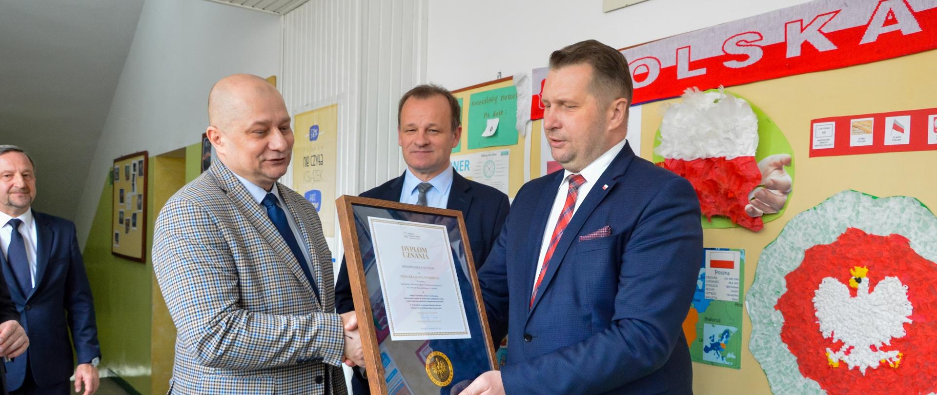 W szkolnym korytarzu z białymi ścianami stoi minister Czarnek, który wręcza mężczyźnie w jasnoszarej marynarce oprawiony w ramki dokument, za nimi stoi mężczyzna w czarnym garniturze, dalej na ścianie korkowa tablica do której przypięty jest biało-czerwony szalik z napisem Polska.