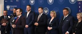 Pod białą ścianą przy której stoją niebieskie banery stoi grupa elegancko ubranych ludzi, jeden mężczyzna mówi do mikrofonu na stojaku.