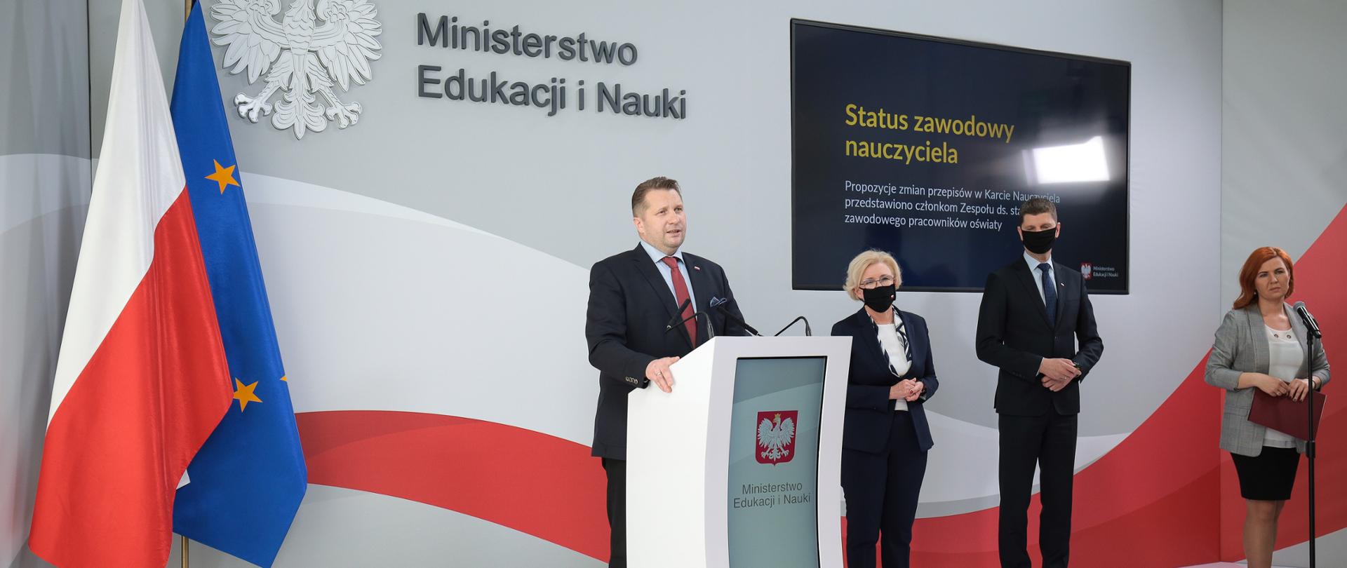 Minister Czarnek przemawia zza mównicy, w dalszym planie stoją wiceministrowie edukacji oraz rzecznik prasowy. W tle ścianka z napisem Ministerstwo Edukacji i Nauki oraz flagi polski i unii