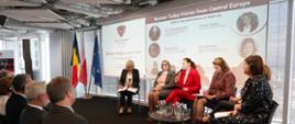 Wokół stolika siedzą paneliści, publiczność patrzy w stronę sceny, za panalistkami wyświetlana jest prezentacja