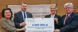 Minister Czarnek, mężczyzna w garniturze, kobieta w czarnej marynarce i kobieta w marynarce w czarno-białą kratkę stoją i trzymają razem wielki symboliczny czek z napisem 3 000 000 zł.