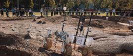 Czterokołowy robot jedzie po nierównym piaszczystym podłożu, w tle za barierką z taśmy stoi grupa ludzi.