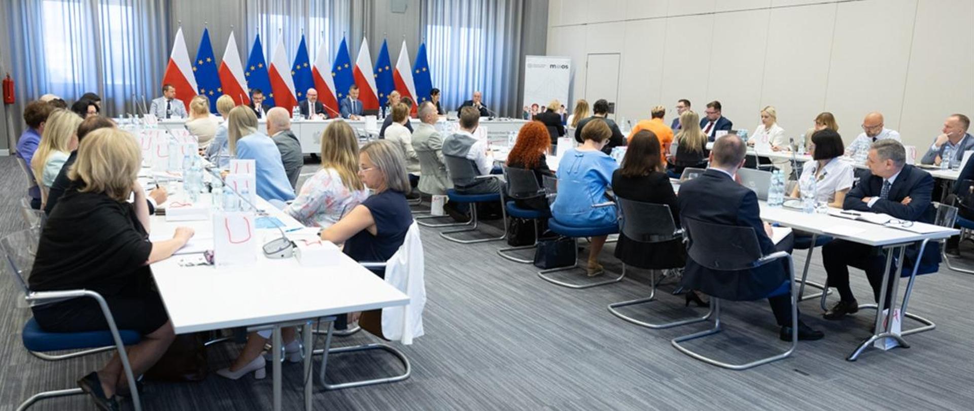 Grupa osób siedzi przy stole ustawionym w podkowę. Za stołem prezydialnym flagi biało-czerwone i unijne 