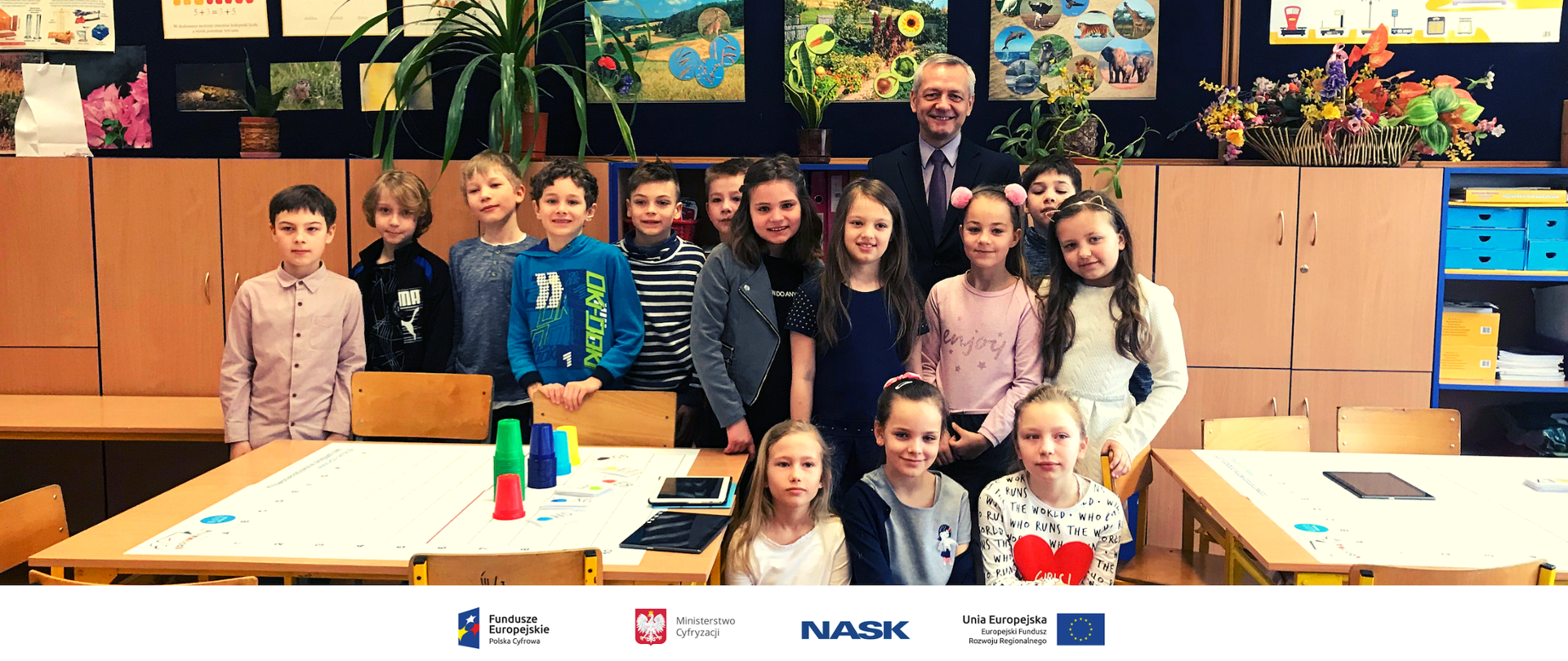 Minister Marek Zagórski w grupie dzieci. W tle ściana z pracami plastycznymi dzieci.