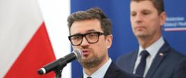 Mężczyzna w garniturze i okularach mówi do mikrofonu, za nim stoi wiceminister Piontkowski, dalej niebieska ściana, z boku polska flaga.
