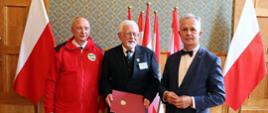 W sali z wyłożonymi drewnianymi panelami ścianami stoi trzech mężczyzn - dwóch w garniturach, jeden w czerwonej kurtce, za nimi pod ścianą polskie flagi.