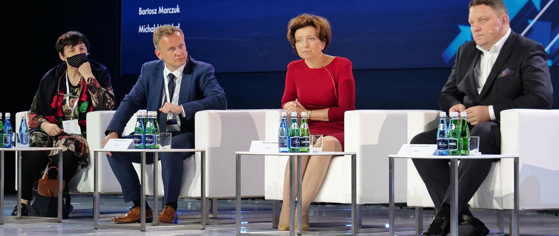 W sali siedzą cztery osoby. W środku siedzi minister Maląg ubrana w czerwoną sukienkę. Obok stolik z butelkami z wodą mineralną.