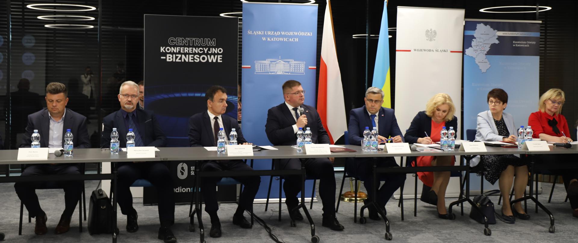 Grupa osób siedzi przy stole. W środku wiceminister Rzymkowski mówi do mikrofonu. W tle banery oraz flagi - polska i ukraińska. 