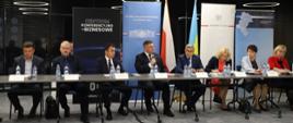 Grupa osób siedzi przy stole. W środku wiceminister Rzymkowski mówi do mikrofonu. W tle banery oraz flagi - polska i ukraińska. 