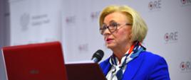 Minister Machałek w niebieskim ubraniu stoi i mówi do mikrofonu, za nią ściana z napisami ORE.