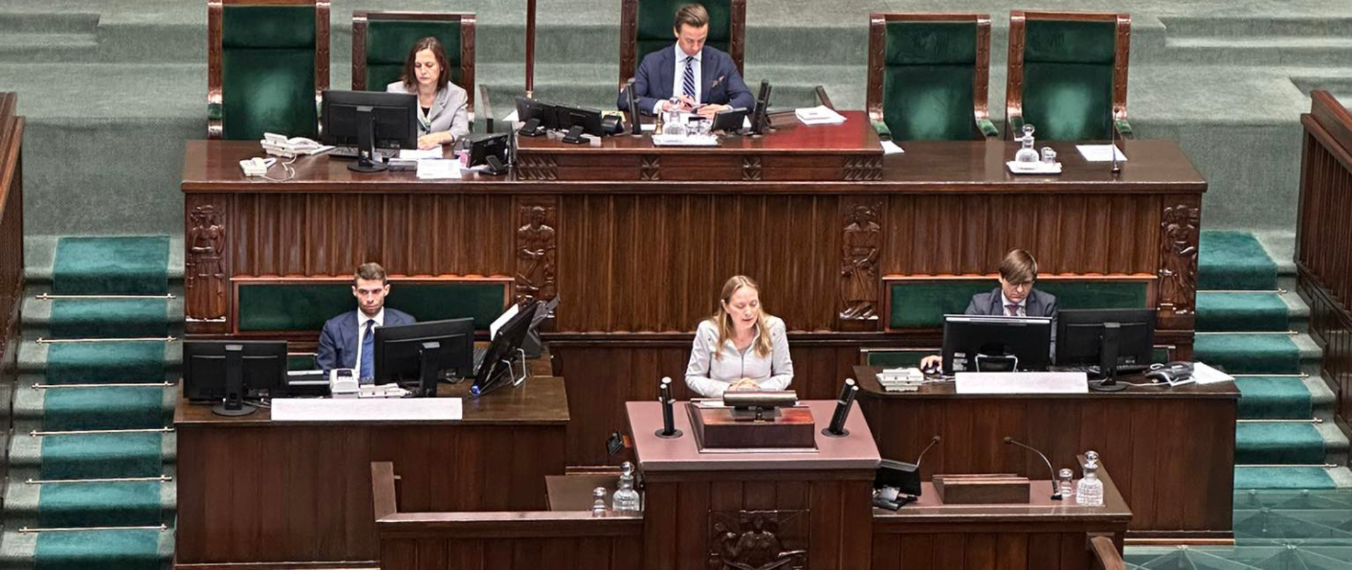 Minister Katarzyna Pełczyńska-Nałęcz w sejmowej mównicy. Za nią siedzi wicemarszałek i inne osoby z obsługi posiedzenia.