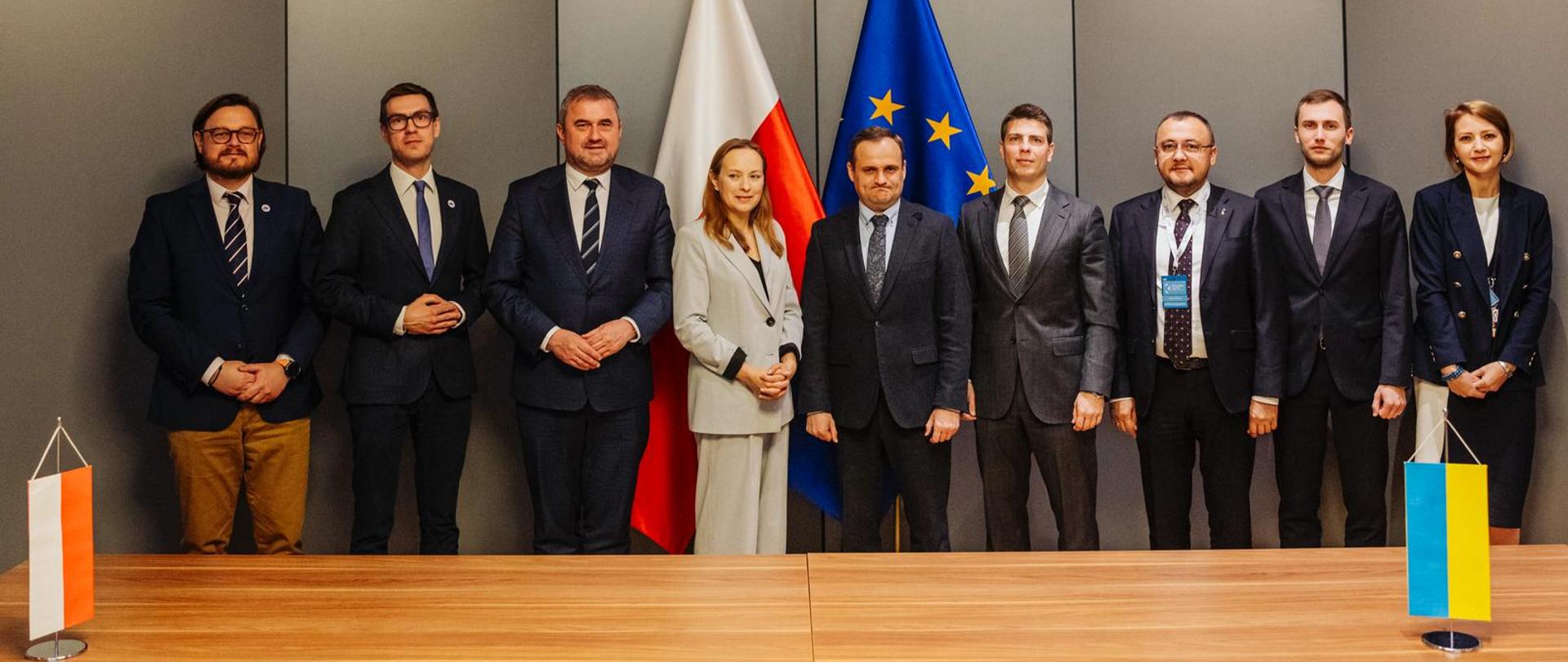 Grupa osób stoi obok siebie przed drewnianym stołem. Wśród nich w środku stoi minister Katarzyna Pełczyńska-Nałęcz.