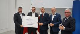 Pod białą ścianą stoi pięciu mężczyzn w garniturach, dwóch z lewej trzyma wielki symboliczny czek z napisem Branżowe Centrum Umiejętności - 9 889 300 zł.