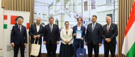 Pod ścianą z pionowych drewnianych deseczek, na której wisi wielki ekran na którym widać zdjęcie starego budynku, stoi grupa siedmiu osób - pięciu mężczyzn w garniturach, i dwie kobiety, obok nich węgierska flaga.