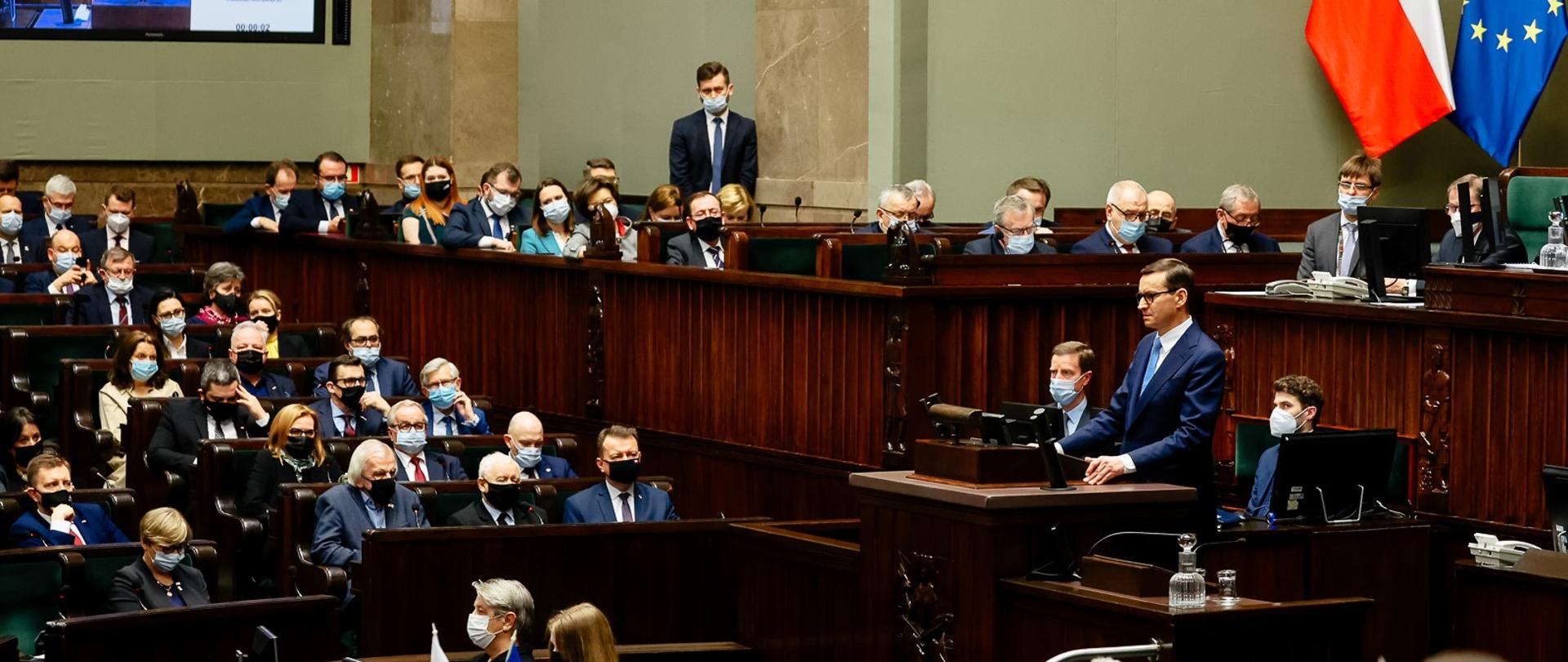 Premier przemawia na mównicy sejmowej, w tle siedzą posłowie.