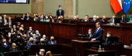 Premier przemawia na mównicy sejmowej, w tle siedzą posłowie.