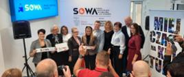 Pod ścianą z napisem SOWA stoi duża grupa ludzi, kilkoro trzyma kawałki wstęgi z napisem SOWA.