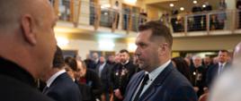 Minister Czarnek ściska rękę komuś poza kadrem, w tle duża sala wypełniona ludźmi, wielu ma czarne galowe mundury.