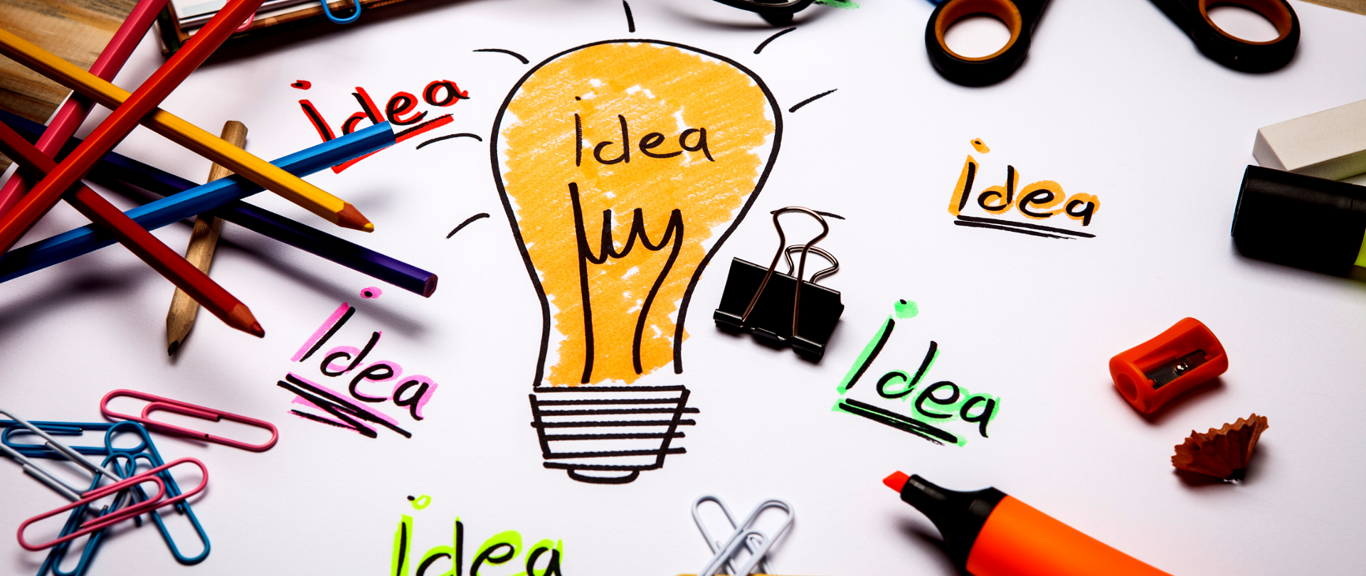 Zdjęcie - biała kartka papieru, na środku rysunek żółtej żarówki, wokół różnokolorowe napisy "idea", na kartce leżą kolorowe kredki, kolorowe spinacze, flamastry.