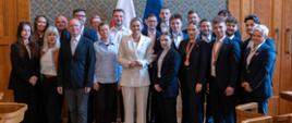 Duża grupa osób, zarówno mężczyzn, jak i kobiet, w różnym wieku, pozuje do zdjęcia w sali z drewnianymi panelami i tapetą. Kobieta w białym garniturze stoi na środku. W tle widoczne są polskie i europejskie flagi.