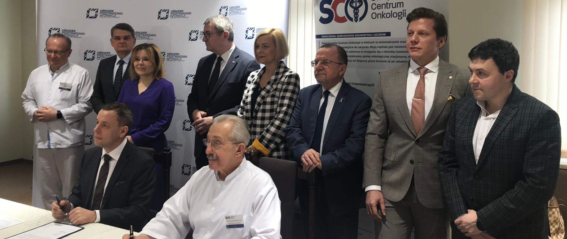 Grupa ludzi stoi za białym stołem przy którym siedzą mężczyzna w garniturze i mężczyzna w białej koszuli, którzy podpisują leżące dokumenty.