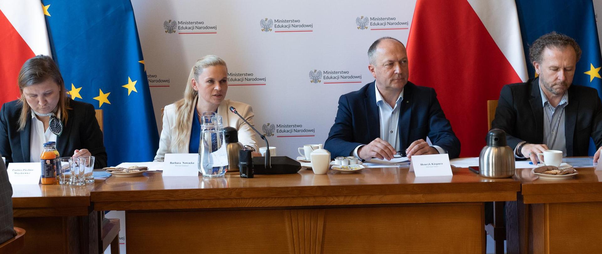 Na tle białej ścianki z napisami Ministerstwo Edukacji Narodowej oraz flag Polski i UE siedzą przy drewnianym stole od lewej: wiceminister Paulina Piechna-Więckiewicz, minister Barbara Nowacka, wiceminister Henryk Kiepura oraz dyrektor generalny Robert Bartold.