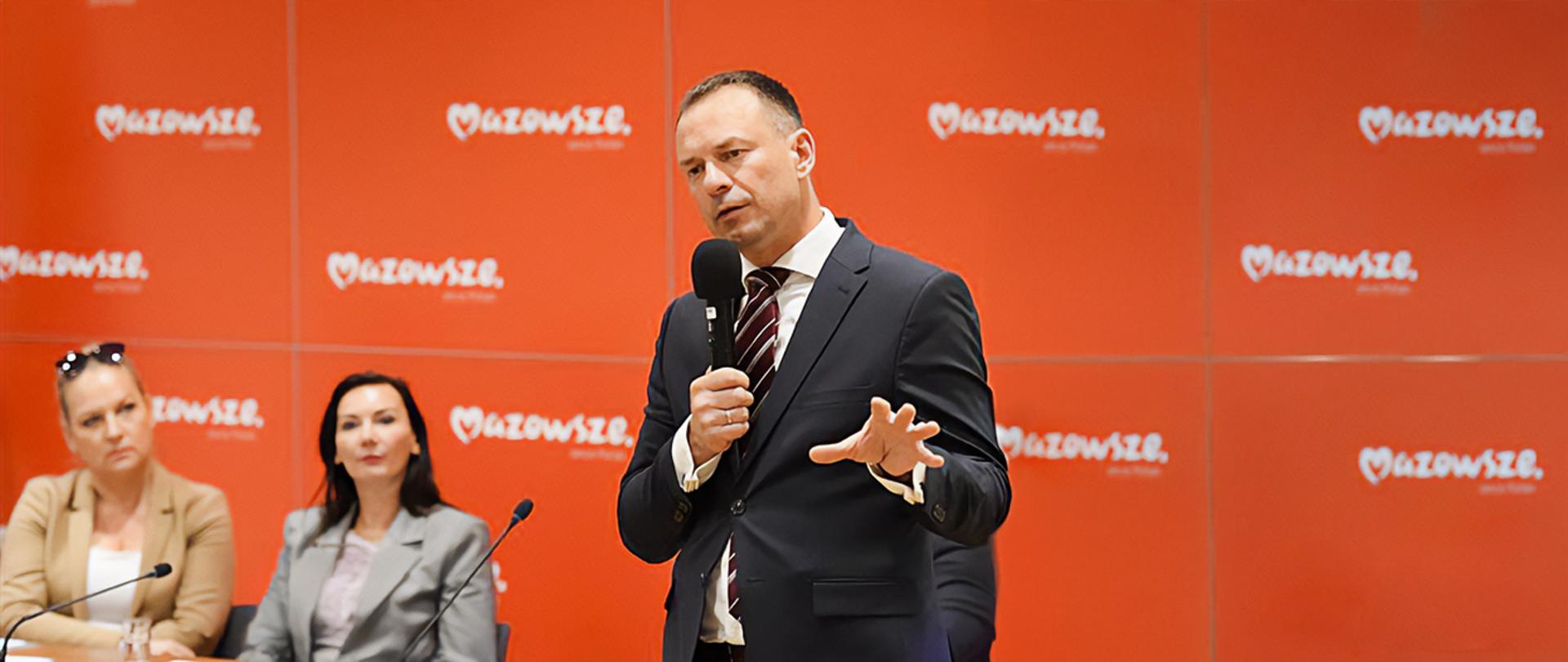 Piotr Borys na konferencji w Urzędzie Marszałkowskim Województwa Mazowieckiego. W ręku trzyma mikrofon, za nim czerwona ścianka z logiem województwa
