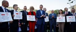 Premier stoi wśród ludzi trzymających czeki.