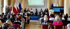Na środku sali ustawiona jest scena, na której siedzi grono panelistów. Jednym z nich jest przemawiająca wiceminister Paulina Piechna-Więckieiwcz, która trzyma w ręku mikrofon. Za panelistami ustawione są flagi Polski i Unii Europejskiej, a nad nimi wisi ekran, na którym wyświetlane są podstawowe informacje o trwającym panelu. Przed sceną ustawionych jest kilka rzędów krzeseł, na których siedzą uczestnicy konferencji. 
