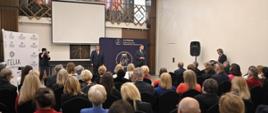 Widok z tyłu na wypełnioną ludźmi salę, z przodu stoi mężczyzna w garniturze i mówi do mikrofonu na stojaku, po obu jego stronach stoi dwóch mężczyzn, za nimi ciemnoszary baner, dalej wielki ekran, w tle widać biegnący dookoła ścian sali balkon.