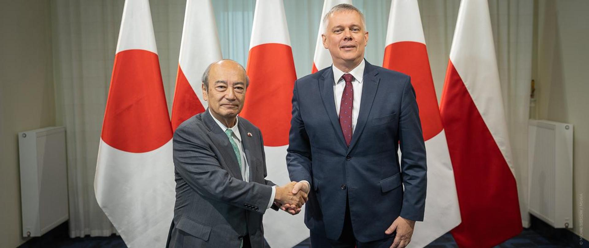 Dwóch mężczyzn w geście powitania na tle flag Polski i Japonii. 