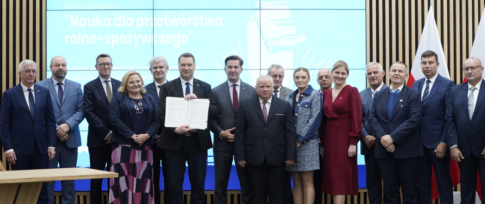 Pod ścianą na której jest wieli ekran stoi duża grupa ludzi, pośrodku minister Czarnek trzyma w ręku białą kartę.
