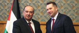 Minister Czarnek ściska rękę mężczyźnie w garniturze, za nimi czarno-biało-zielona flaga a za nią ściana w zielony wzorek.