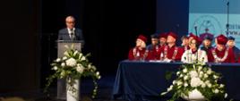Mężczyzna w garniturze i z wąsami stoi przy mównicy i mówi do mikrofonu, obok niebieski stół przy którym siedzą ubrani w czerwone ceremonialne stroje ludzie, przed stołem bukiet białych kwiatów.